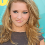 A person with long, voluminous blonde hair styled in loose waves, wearing a dark top and large hoop earrings, posing in front of a teal and yellow promotional backdrop.