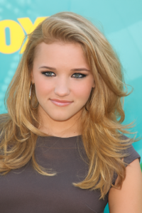 A person with long, voluminous blonde hair styled in loose waves, wearing a dark top and large hoop earrings, posing in front of a teal and yellow promotional backdrop.
