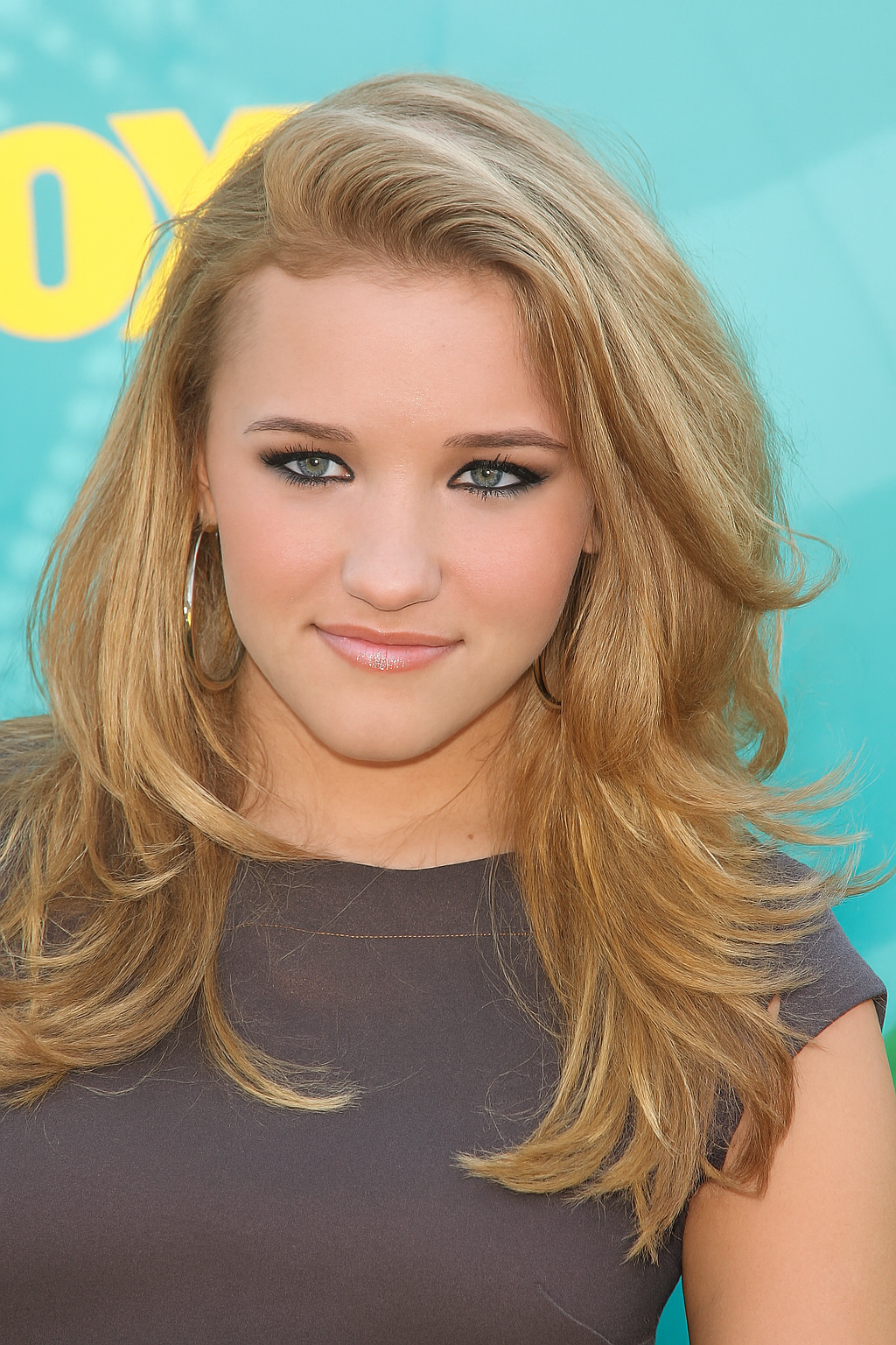 A person with long, voluminous blonde hair styled in loose waves, wearing a dark top and large hoop earrings, posing in front of a teal and yellow promotional backdrop.