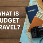 Travel essentials on a wooden table including a passport, map, wallet with dollar bills, camera, and straw hat — representing budget travel.
